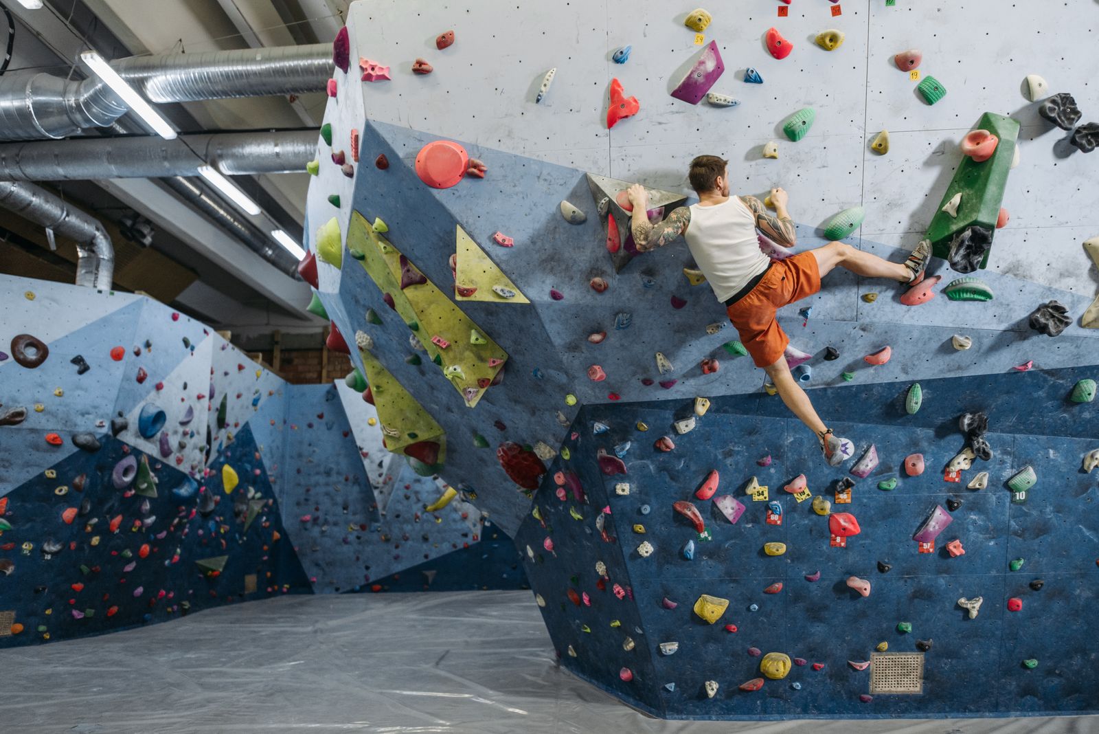 A climber in an orange shorts stretching across a steep blue bouldering wall covered in colorful holds