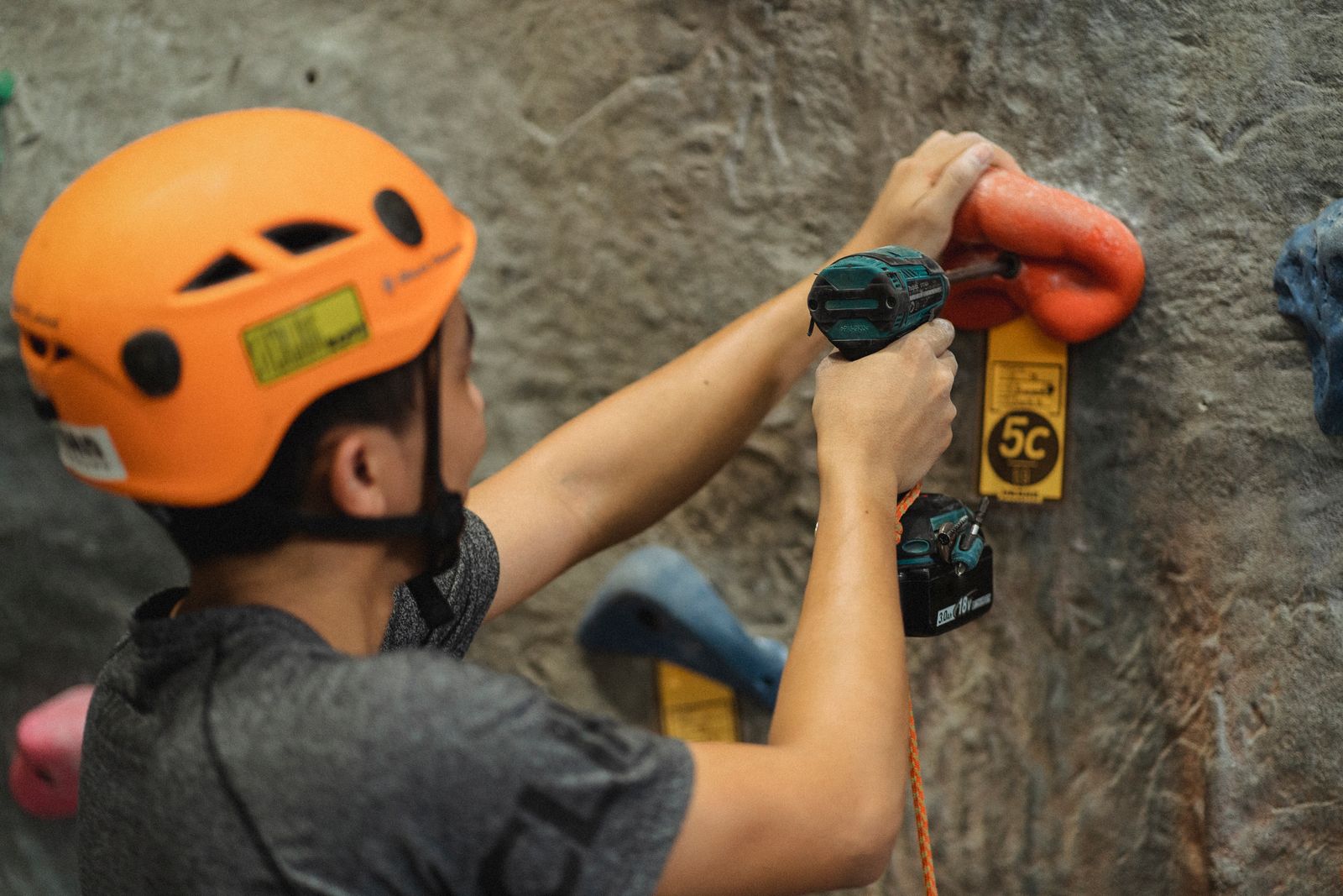A route setter in an orange helmet fastening a climbing hold to a wall with a drill