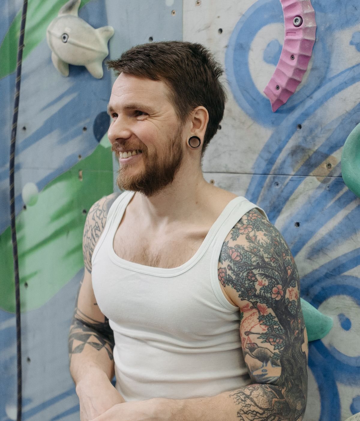 Cole Brennan, a bearded man with arm tattoos in a white tank top, smiling beside a painted climbing wall