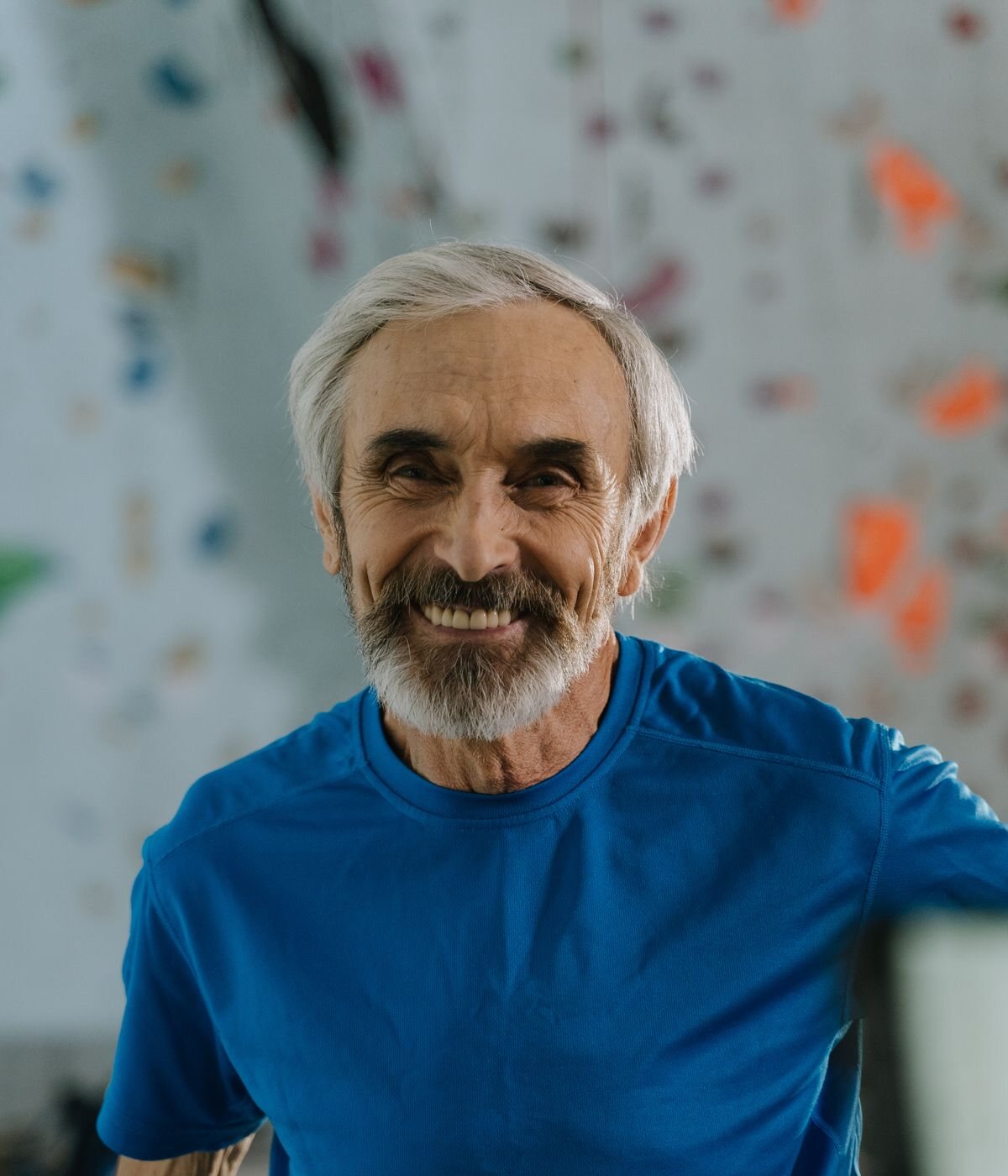 Hank Ostrander, a smiling man with white hair and a short beard in a blue t-shirt, in front of a climbing wall