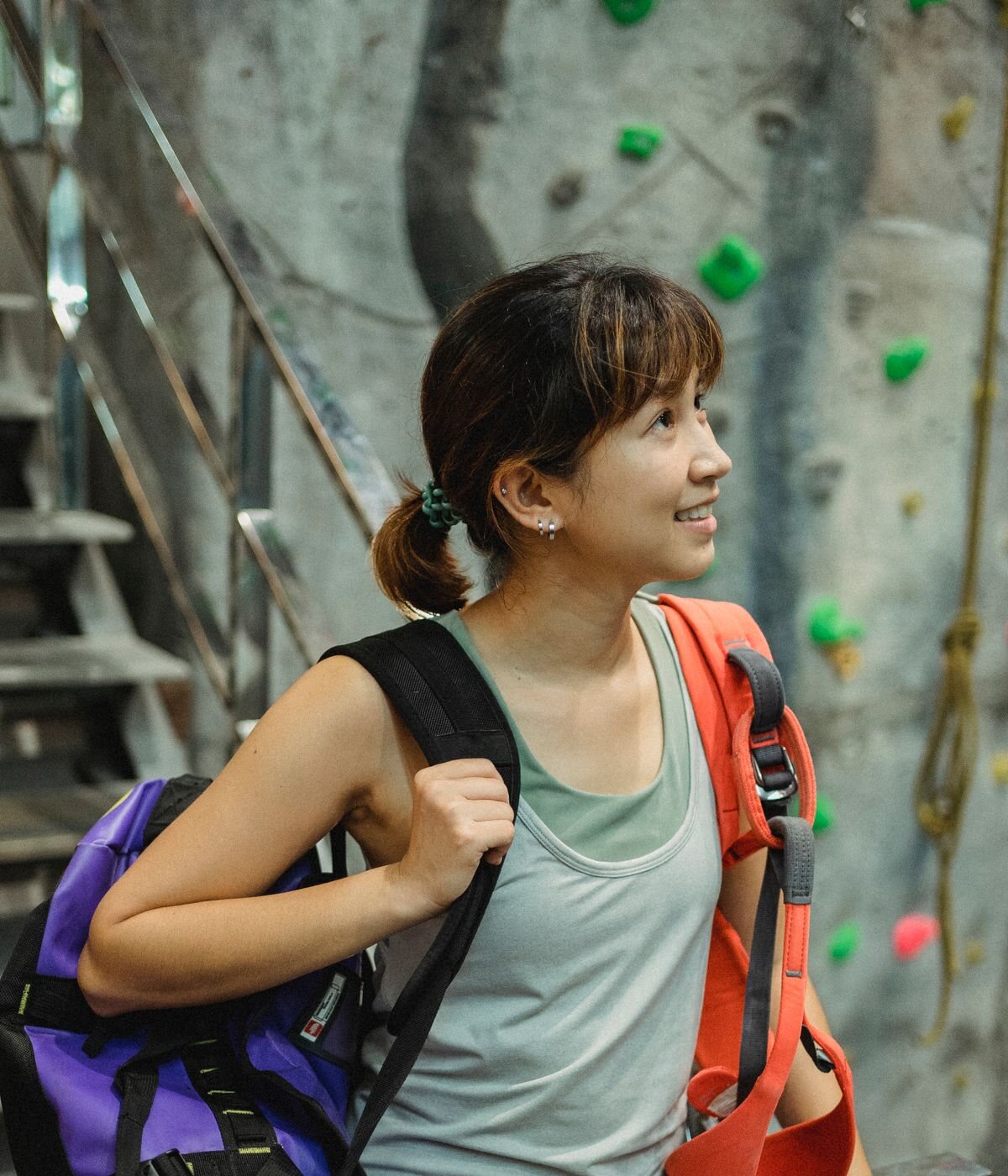 Naomi Ito, smiling and looking up, with a backpack and an orange harness over her shoulder in front of a climbing wall